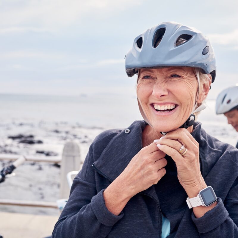 Older woman wearing a muted blue helmet. She's smiling as she is buckling it up underneath her chin.